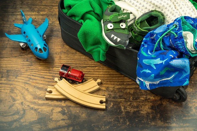 An open suitcase on a floor with kids' stuff strewn about, both in and outside of it: a toy plane, wood train track pieces and a toy train, shoes with a crocodile design, swim shorts, and more.