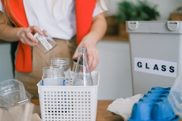 A woman recycling indoors, in her kitchen. She's putting glassware into a basket, next to an indoor recycling bin with "GLASS" written on it.
